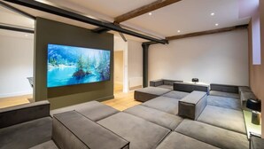 A modern living room with a large flat-screen TV mounted on a wall, grey cushioned seating, exposed wooden beams, and recessed ceiling lighting.