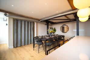 Et moderne spiseområde med et langt bord dækket til seks personer, omgivet af stole. Der er et stort vindue med lodrette skodder, et rundt spejl på væggen og synligt ventilationsrør i loftet.