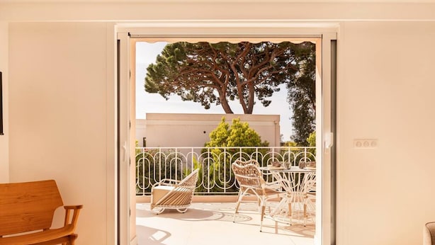 A room with a wooden bench, a sliding glass door leading to an outdoor balcony with white wicker furniture and a tree. - Carla Rosa 7 Appartement Moderne Piscine - 2 BDR (Cannes)