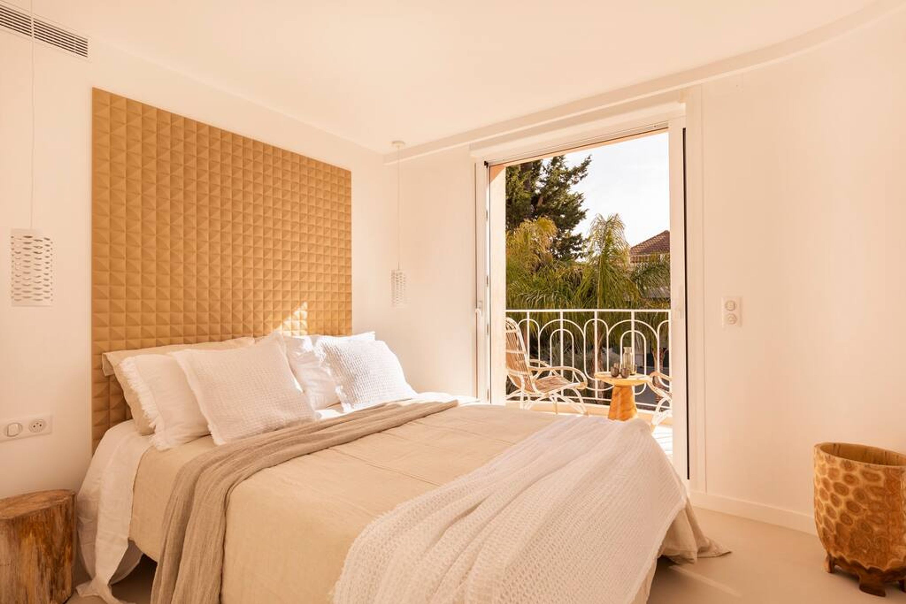 A bedroom with a large bed, a headboard, and a sliding glass door leading to an outdoor area with chairs and a table.