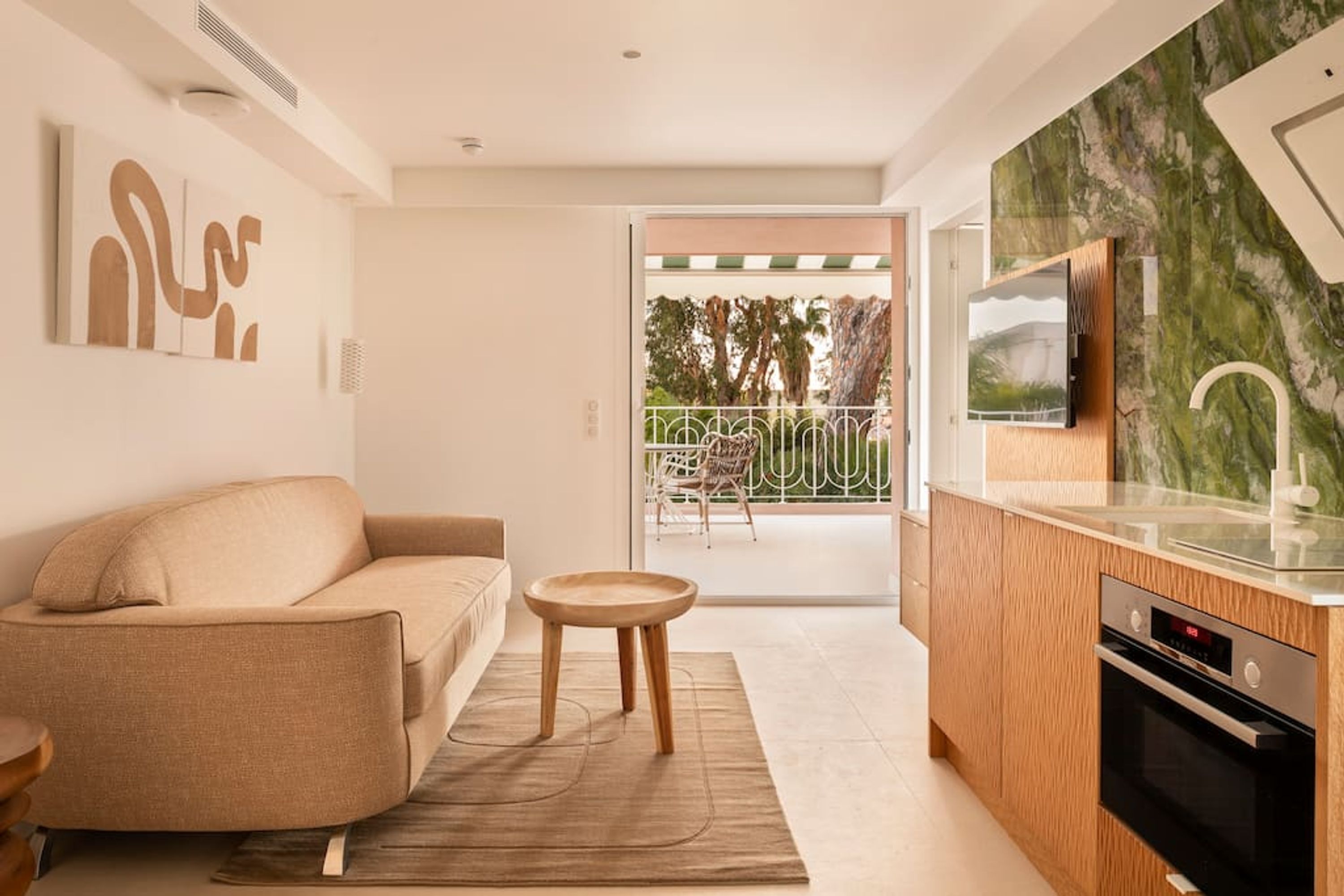 A modern living room with a beige sofa, a small wooden table, and a kitchen area with a marble backsplash and built-in oven.