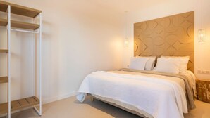 Une chambre à coucher avec un lit, du linge de lit blanc, une tête de lit en bois, une armoire blanche et une table de chevet avec une lampe.