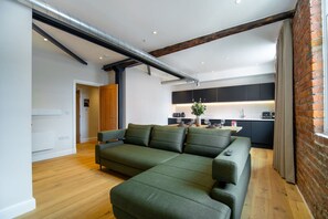 Uma sala de estar moderna com um sofá seccional, uma cozinha ao fundo e paredes de tijolo aparente.