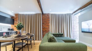 Un salon moderne avec un canapé vert, une table à manger entourée de chaises et un coin cuisine doté d’un mur en briques et de rideaux.