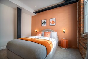 A bedroom with a bed, two wall-mounted lights, a small orange side table, and two framed pictures on the wall.