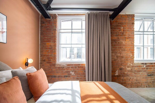 A hotel room with a brick wall, a large window with curtains, a bed with pillows, and a wall-mounted light.