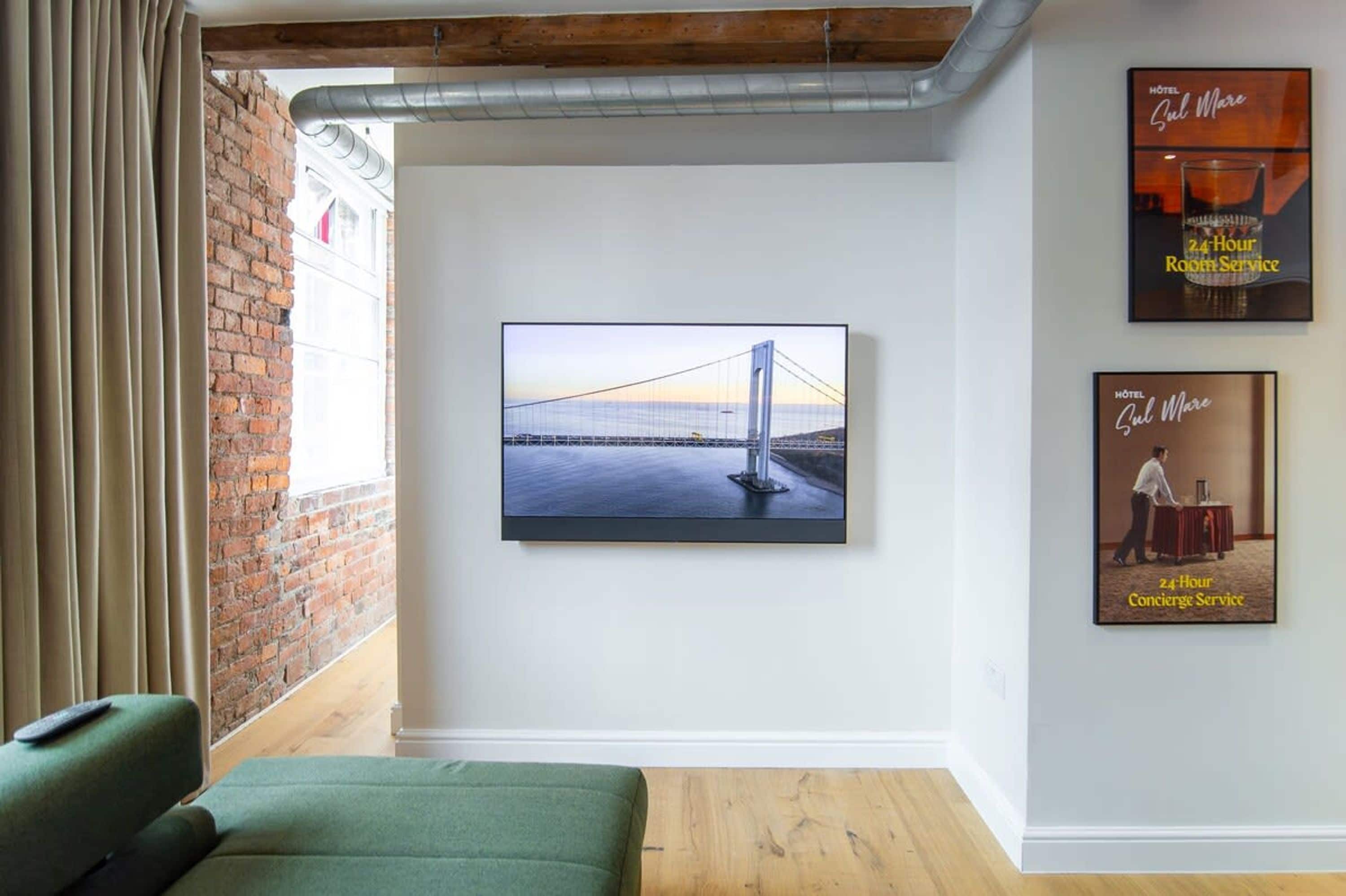 A hotel room with a large window, a green sofa, a flat-screen TV mounted on the wall, and two framed posters advertising 24-hour room service and concierge service.