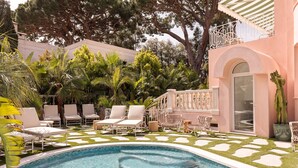 A swimming pool with lounge chairs, surrounded by tropical plants and a pink building with a balcony.