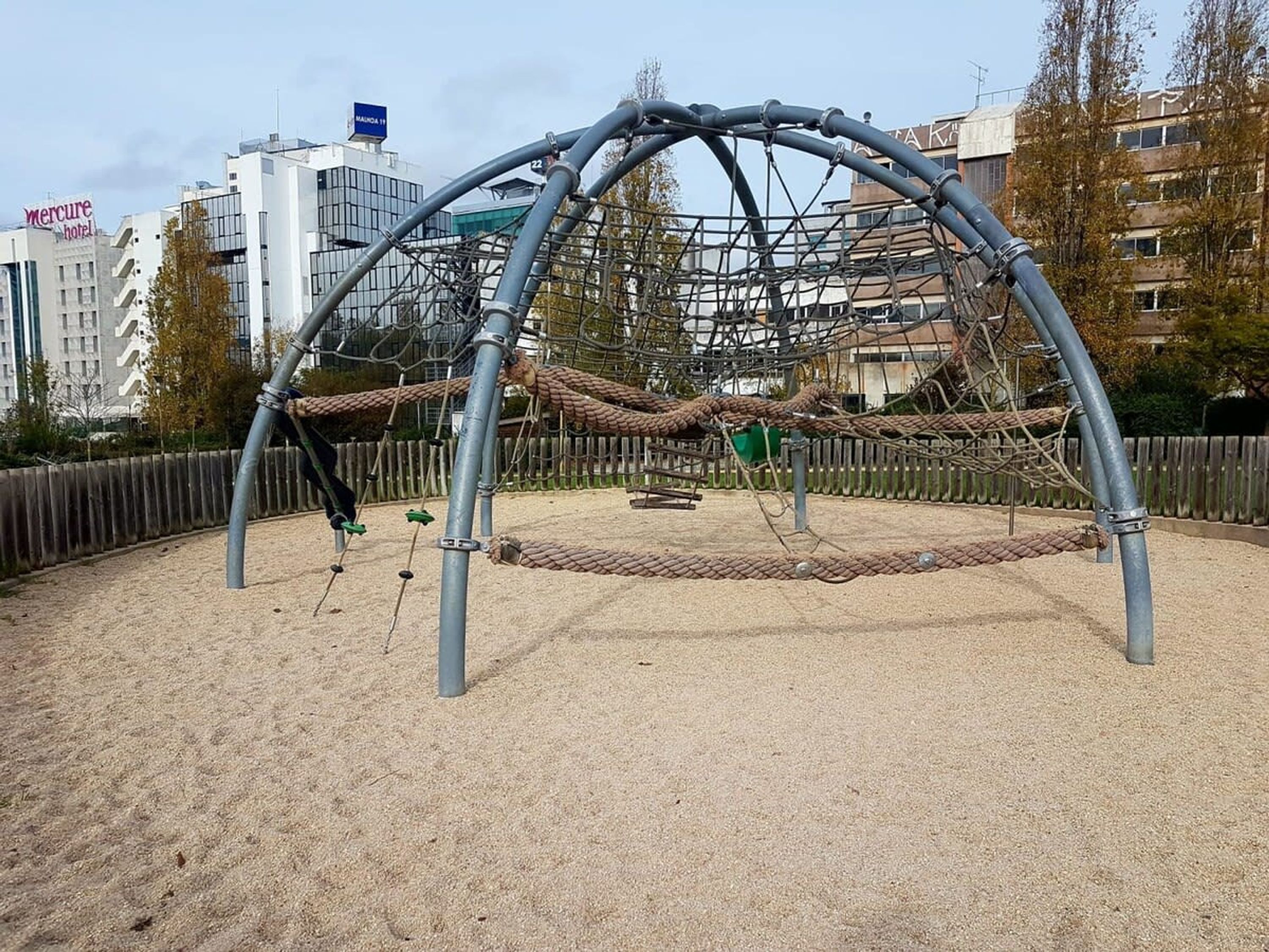 Une aire de jeux avec une structure d’escalade faite de cordes et des balançoires, entourée d’une zone sablonneuse et d’une clôture en bois.