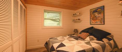 A bedroom with a wooden ceiling, a window, a bed with a patterned quilt, a nightstand, and a wall-mounted shelf with decorative items.
