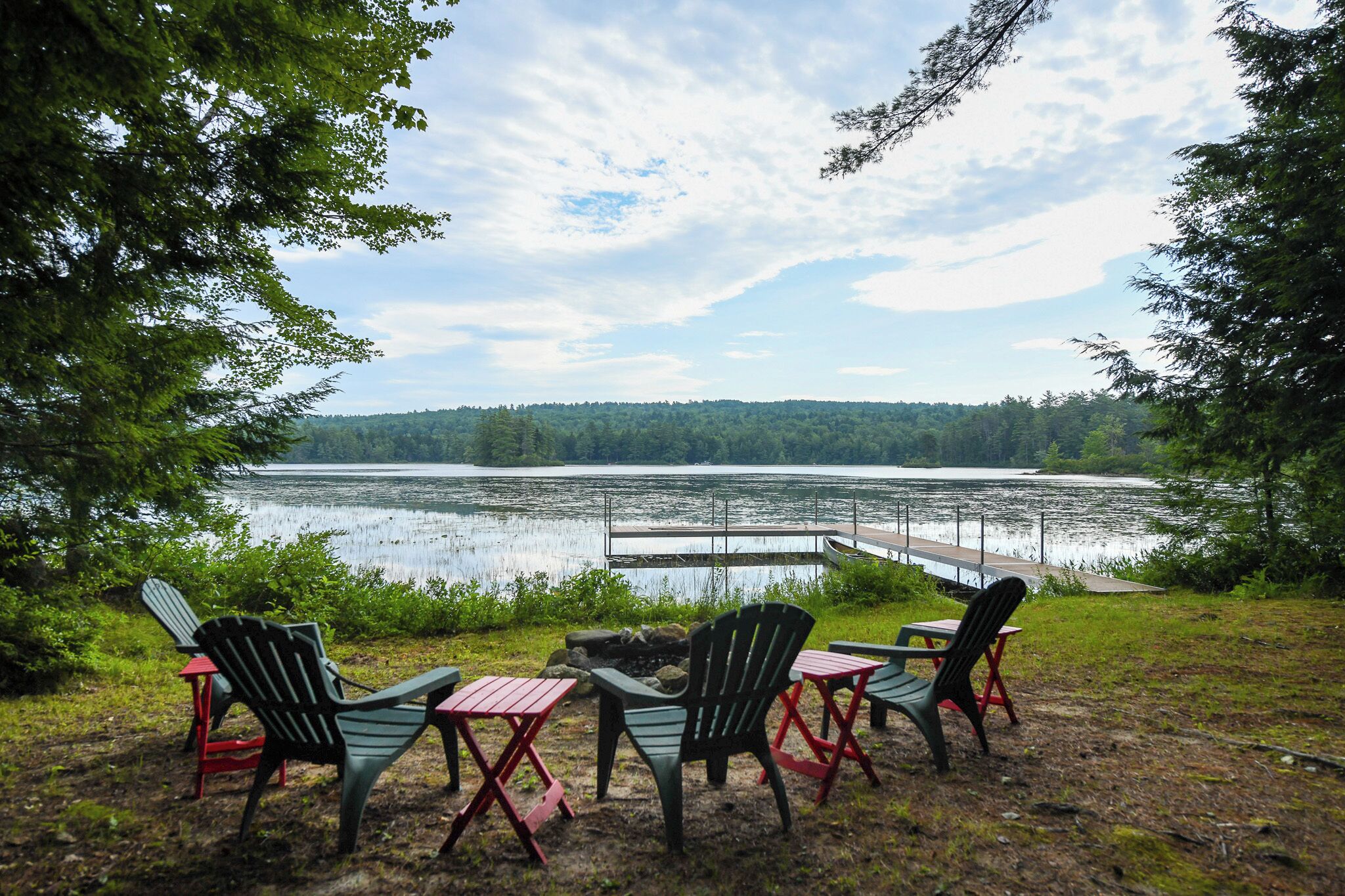 A lakeside seating area with Adirondack chairs and tables, overlooking a calm lake and forested hills.