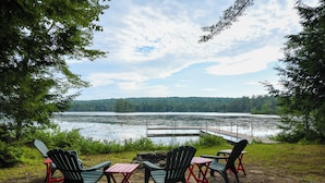 Un espacio de descanso junto al lago con sillas Adirondack y mesas, con vistas a un lago tranquilo y colinas cubiertas de bosque.