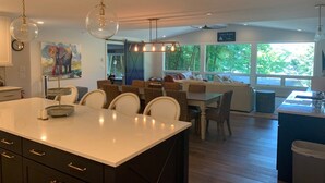 Une cuisine moderne dotée d’un grand îlot central, d’un coin repas avec des meubles en osier et offrant une vue sur l’extérieur grâce à de grandes fenêtres.