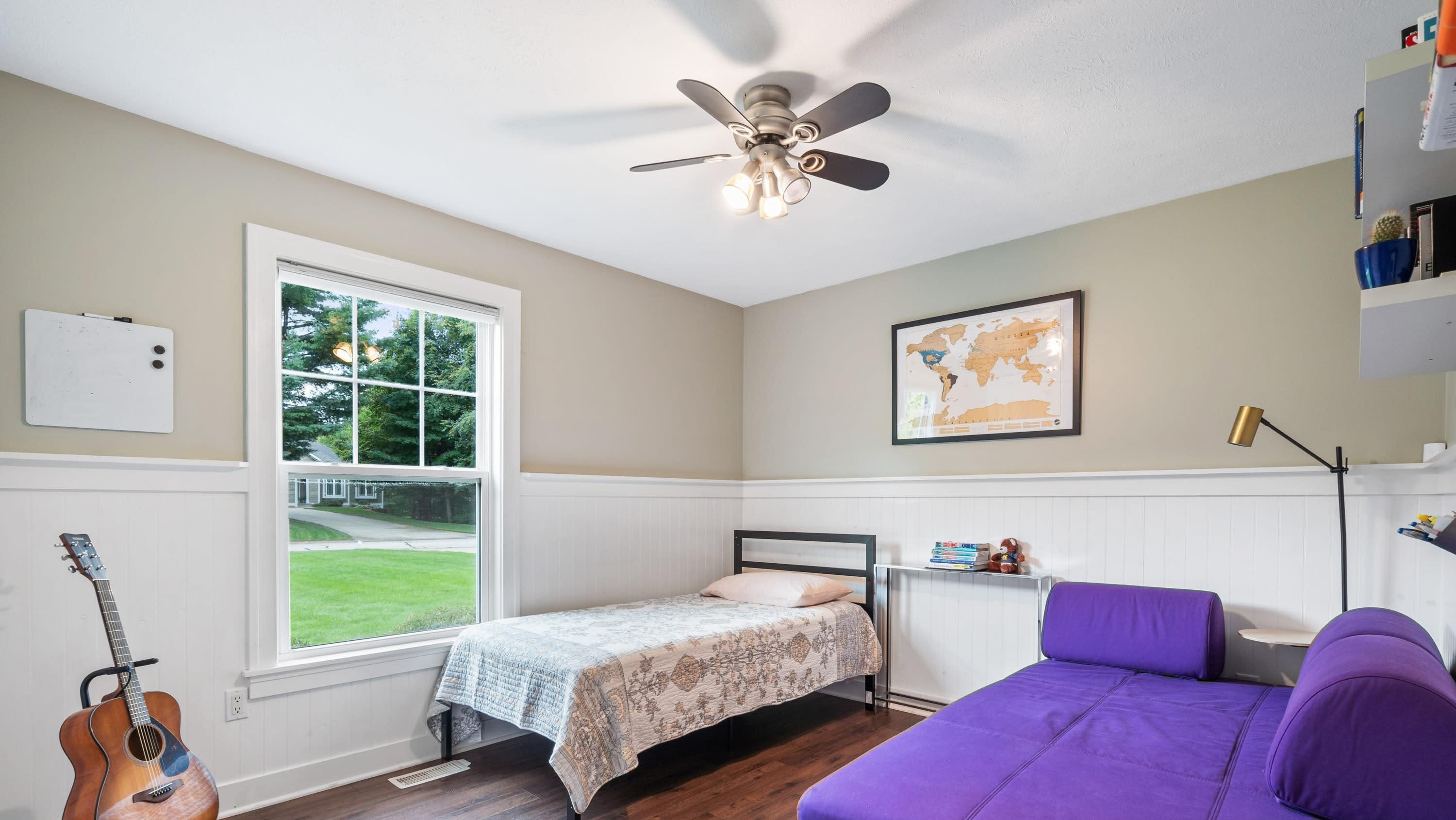 A bedroom with a bed, a guitar, a purple couch, a ceiling fan, a window with a view of a lawn, and a map on the wall.