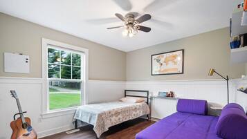 A bedroom with a bed, a guitar, a purple couch, a ceiling fan, a window with a view of a lawn, and a map on the wall.