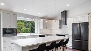 A modern kitchen with white cabinetry, a large island with bar stools, a stainless steel refrigerator, and pendant lighting. - Destined for Relaxation (Saugatuck)