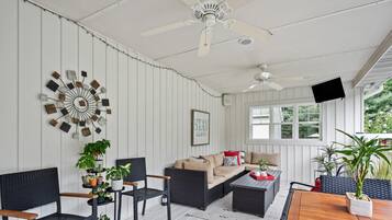 Ein gemütliches Zimmer mit einer Couch, Sesseln, einem Couchtisch und Topfpflanzen. Es gibt einen Deckenventilator, einen Fernseher und ein Fenster mit Blick auf Grünflächen.