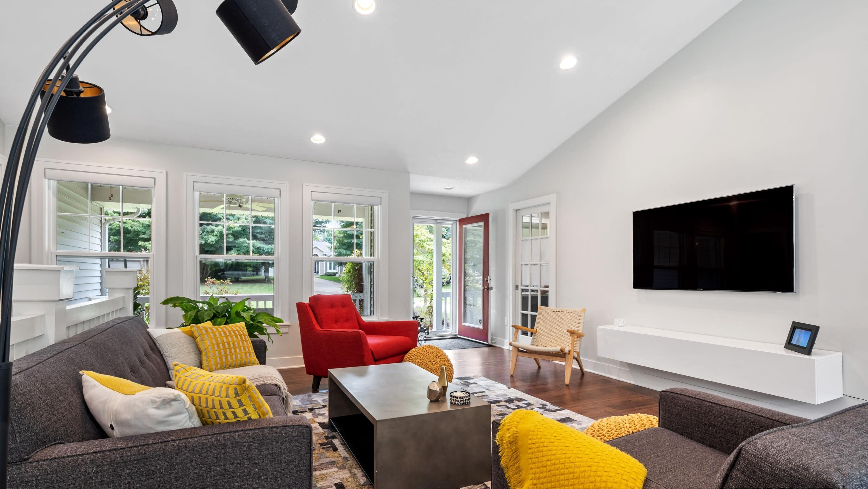 A modern living room with a grey sofa, red armchair, and a coffee table. There is a TV mounted on the wall and a large window with a view of greenery.