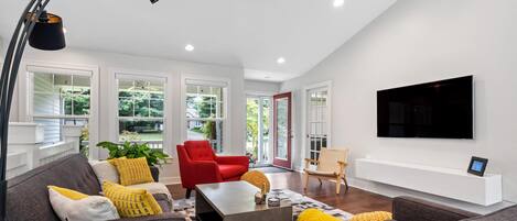 A modern living room with a grey sofa, red armchair, and a coffee table. There is a TV mounted on the wall and a large window with a view of greenery.