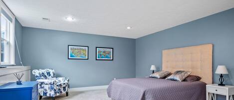 Un dormitorio con cama, mesita de noche, una silla, cómoda y dos cuadros en la pared.