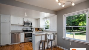 A modern kitchen with white cabinets, stainless steel appliances, and a large window offering a view of greenery. - All Decked Out Farmhouse - Game Room Nearby Beach (Holland)