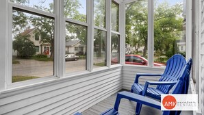 Eine überdachte Veranda mit weißem Holzverkleidung und großen Fenstern, mit blauen Adirondack-Stühlen und Blick auf eine Wohnstraße.
