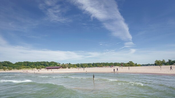 Ein Strand mit blauem Himmel, ruhigem Wasser und sandigem Ufer.