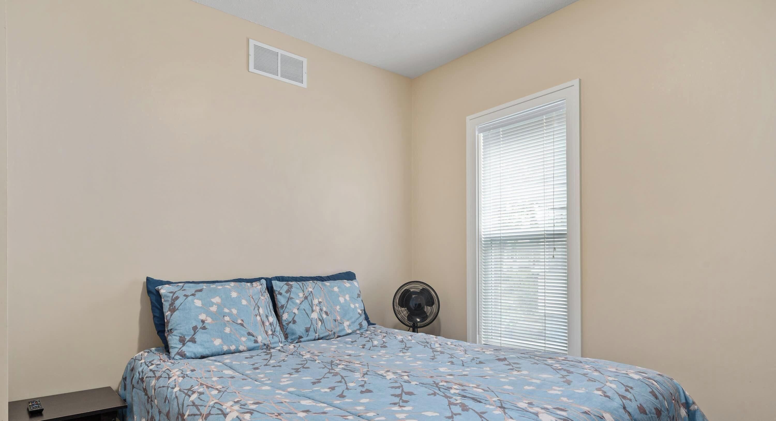 Un dormitorio con cama, mesita de noche, ventilador y ventana con persianas.