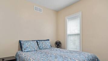 Un dormitorio con cama, mesita de noche, ventilador y ventana con persianas.