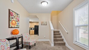 A living room with patterned chairs, a small table, and a decorative orange sphere. A staircase leads to a kitchen area.