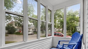 A covered porch with white siding and large windows, featuring blue Adirondack chairs and a view of a residential street.