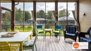 Een overdekt buitengebied met een tafel en stoelen, grote ramen en uitzicht op groen en gebouwen.