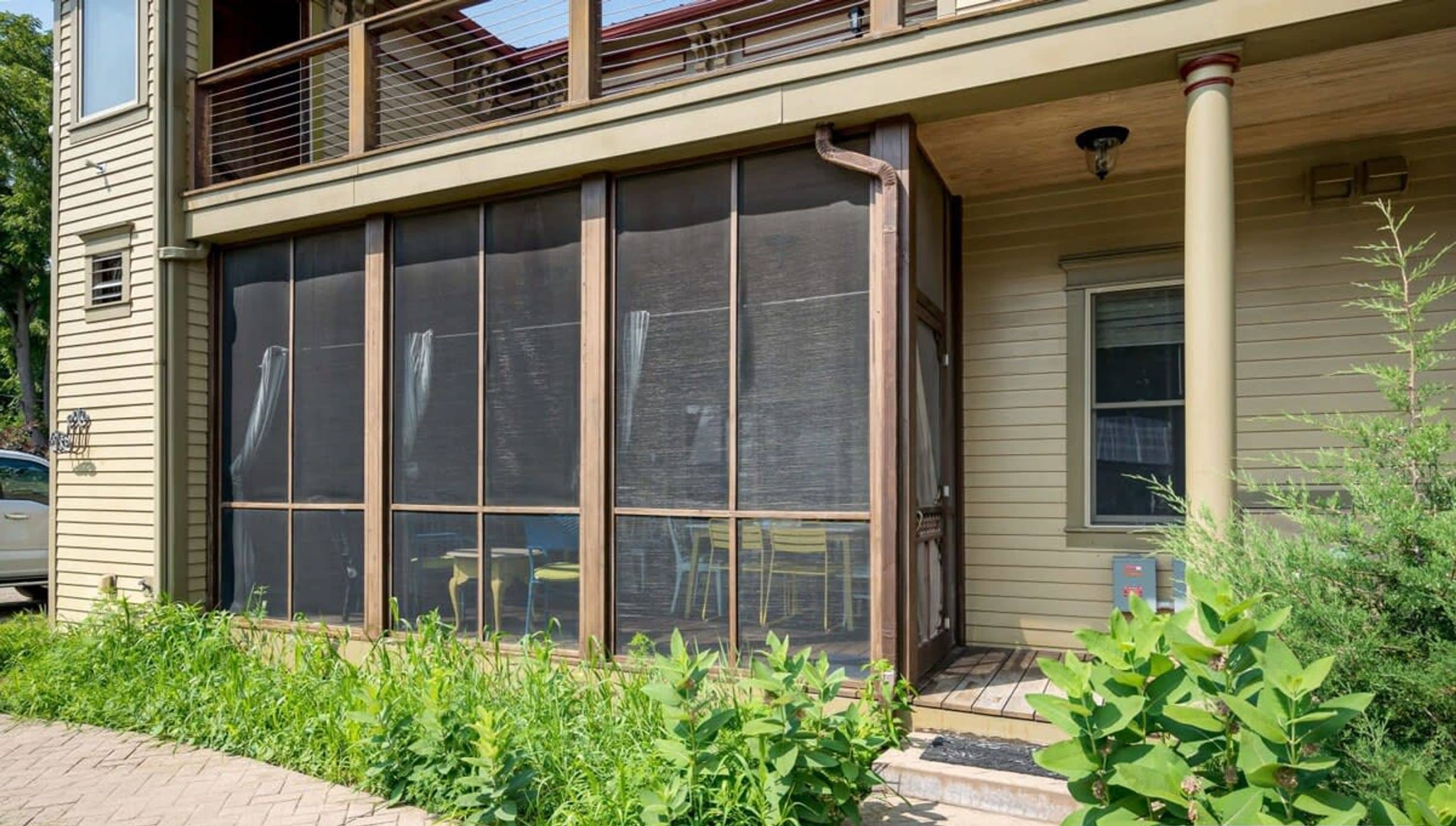 A two-story house with a large screened window on the ground floor, a covered porch, and a small garden area.