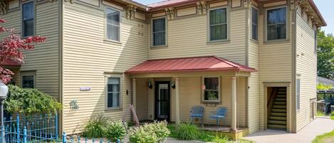 A two-story building with a beige exterior, a red roof, and a front porch with two chairs.