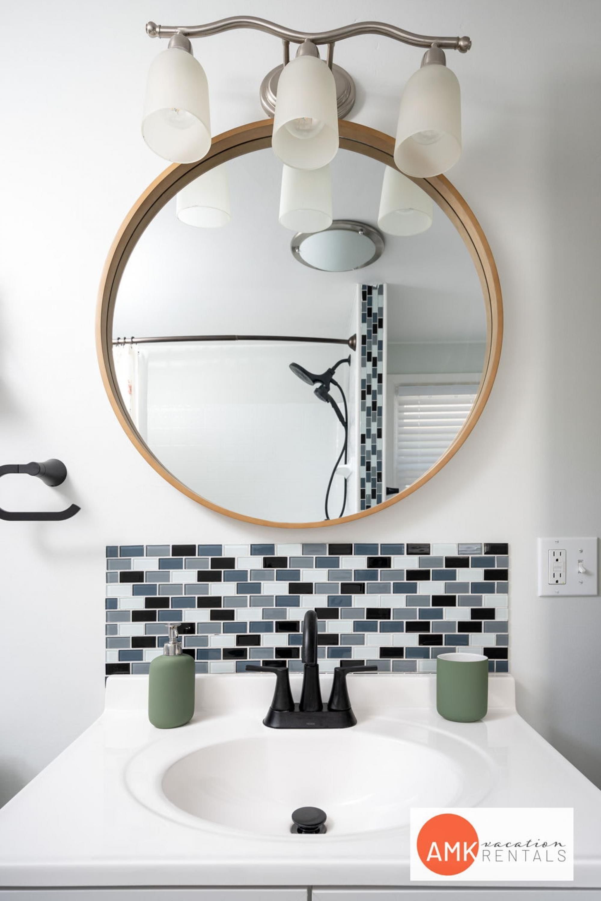 Une salle de bain avec un miroir rond, un lavabo blanc, un robinet noir et un mur recouvert de carreaux.