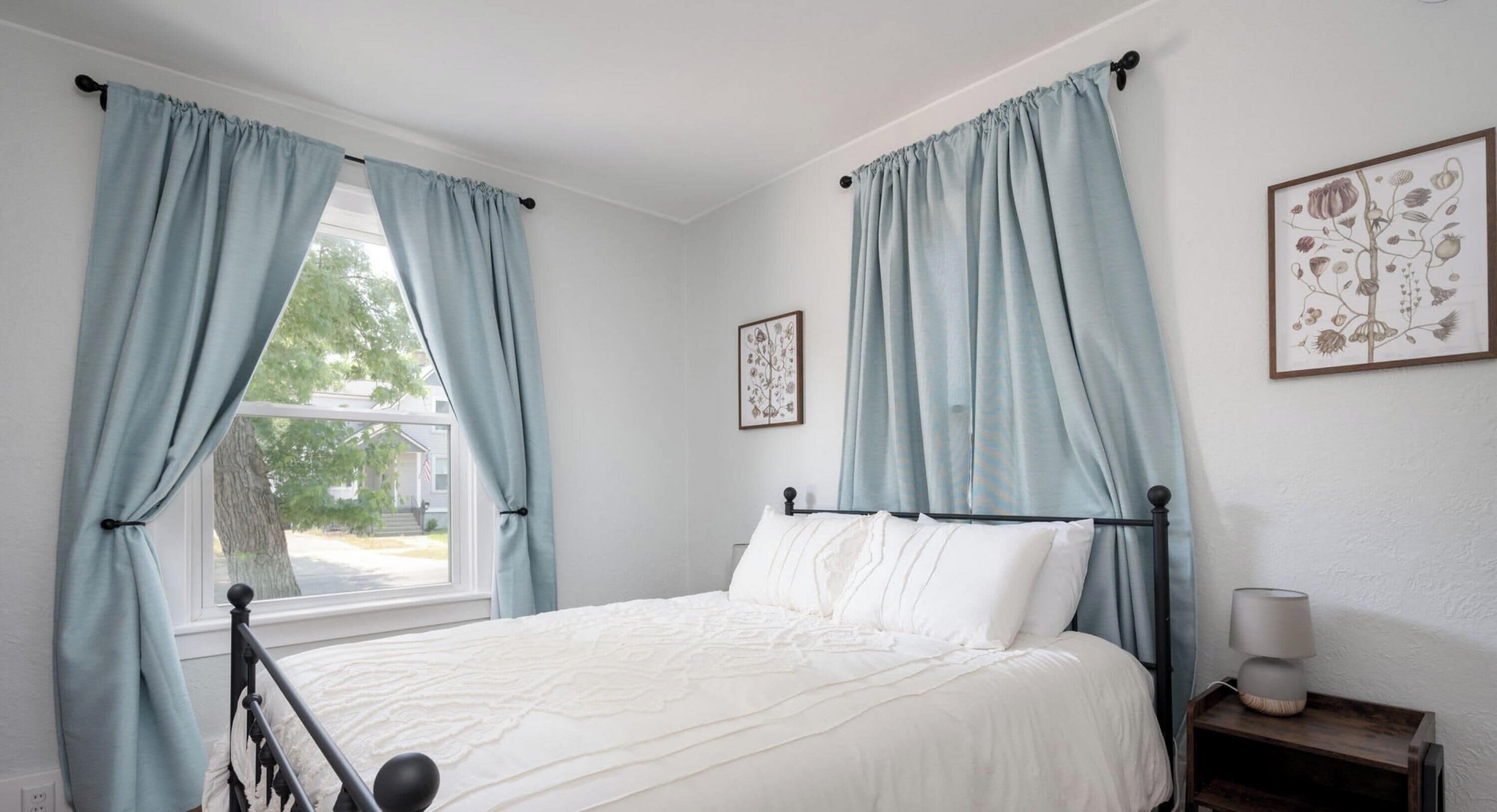 Une chambre à coucher avec un lit, des rideaux, une table de chevet, une lampe et deux reproductions botaniques encadrées accrochées au mur.