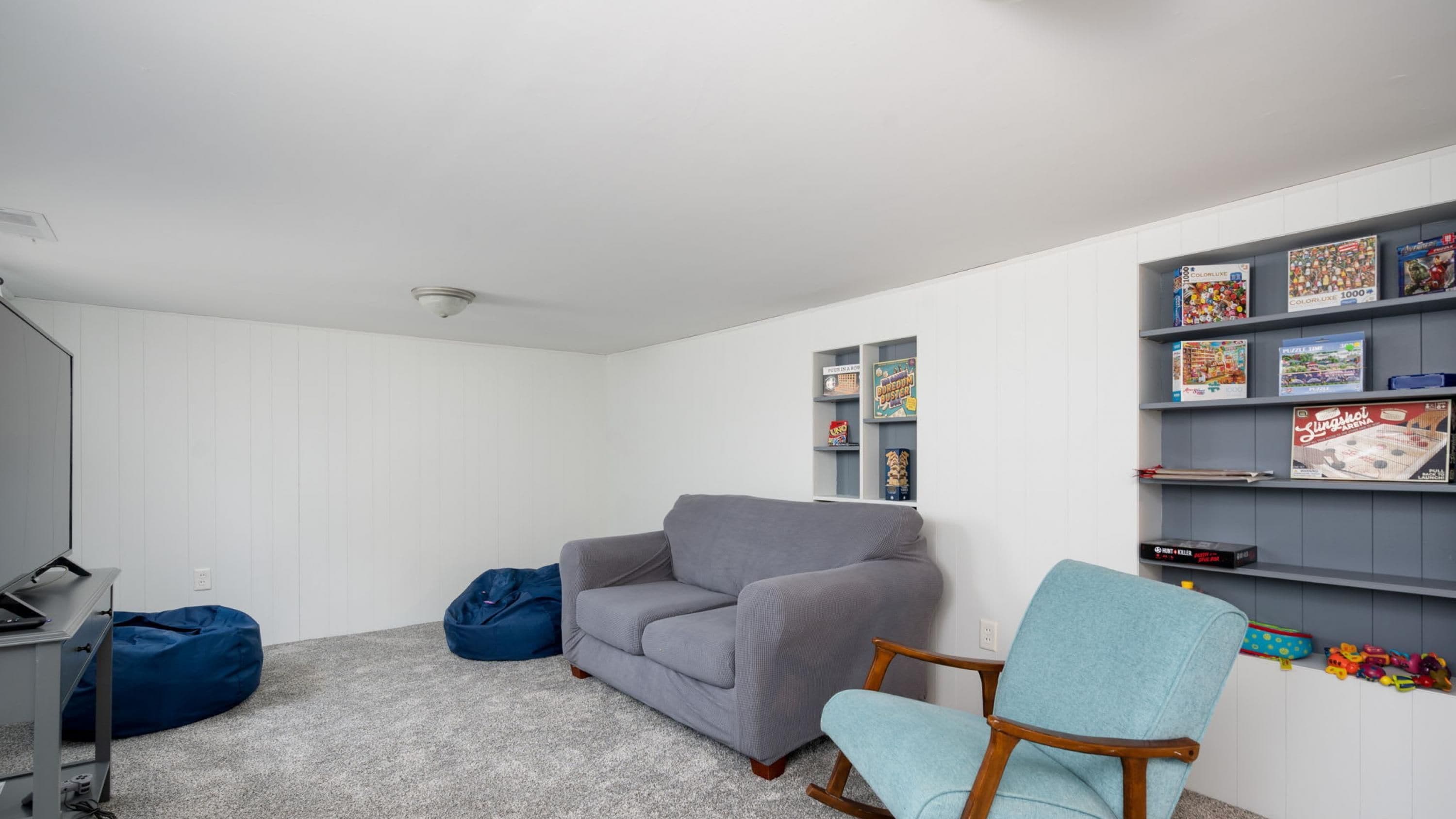 A room with a grey sofa, blue bean bag, and a rocking chair. There are shelves with board games and a desk with a computer.