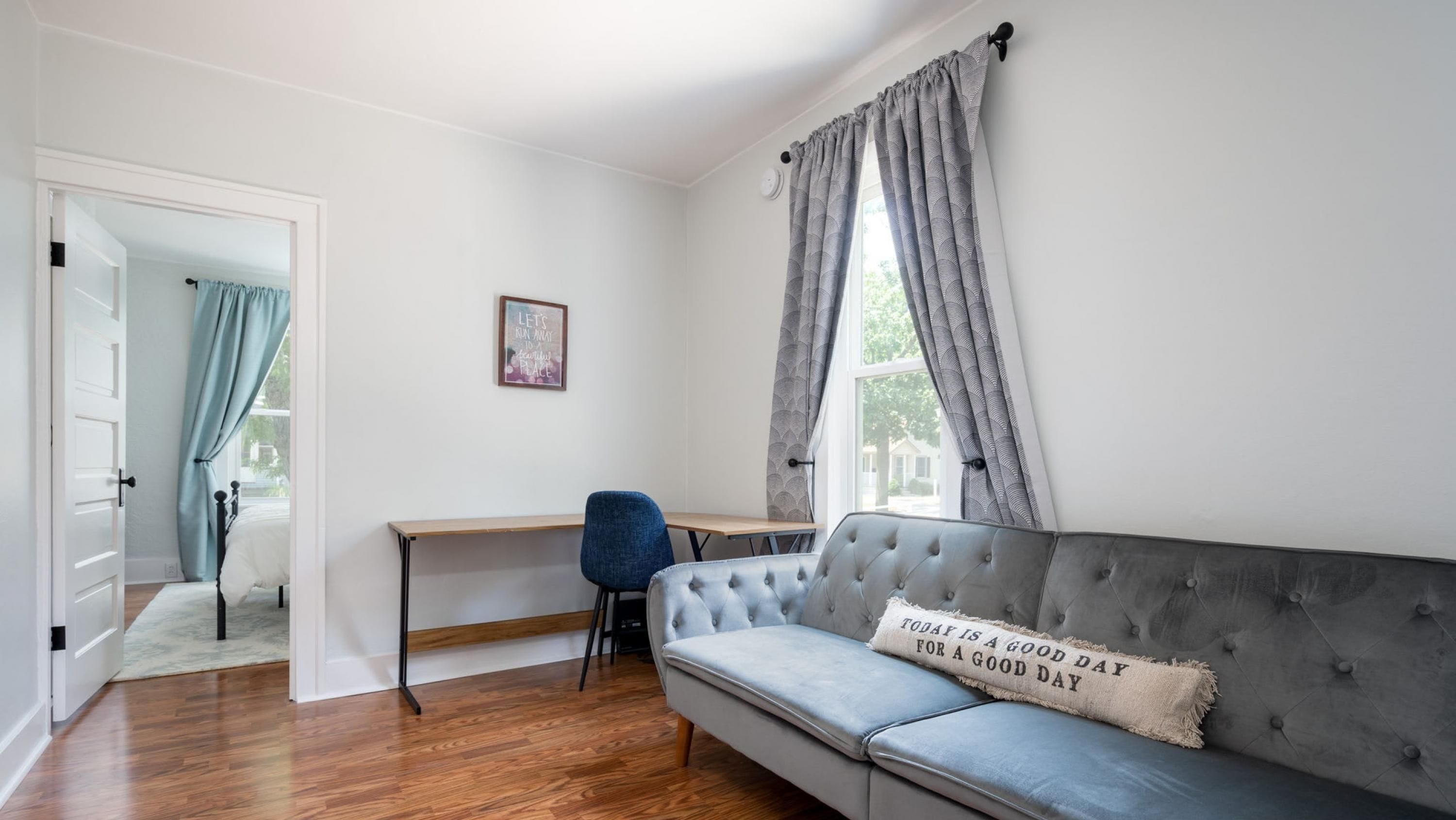 Ein modernes Wohnzimmer mit einem grauen Sofa, Holzboden, einem Schreibtisch und einem Fenster mit Vorhängen.