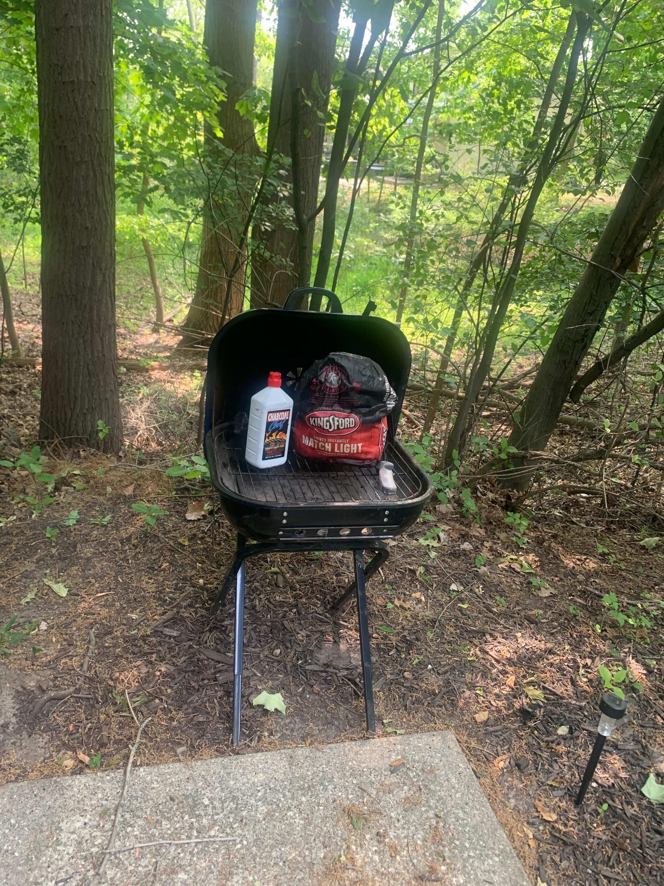 Un barbecue portable à charbon de bois, avec un sac de charbon de bois de la marque Kingsford, situé dans une zone boisée.