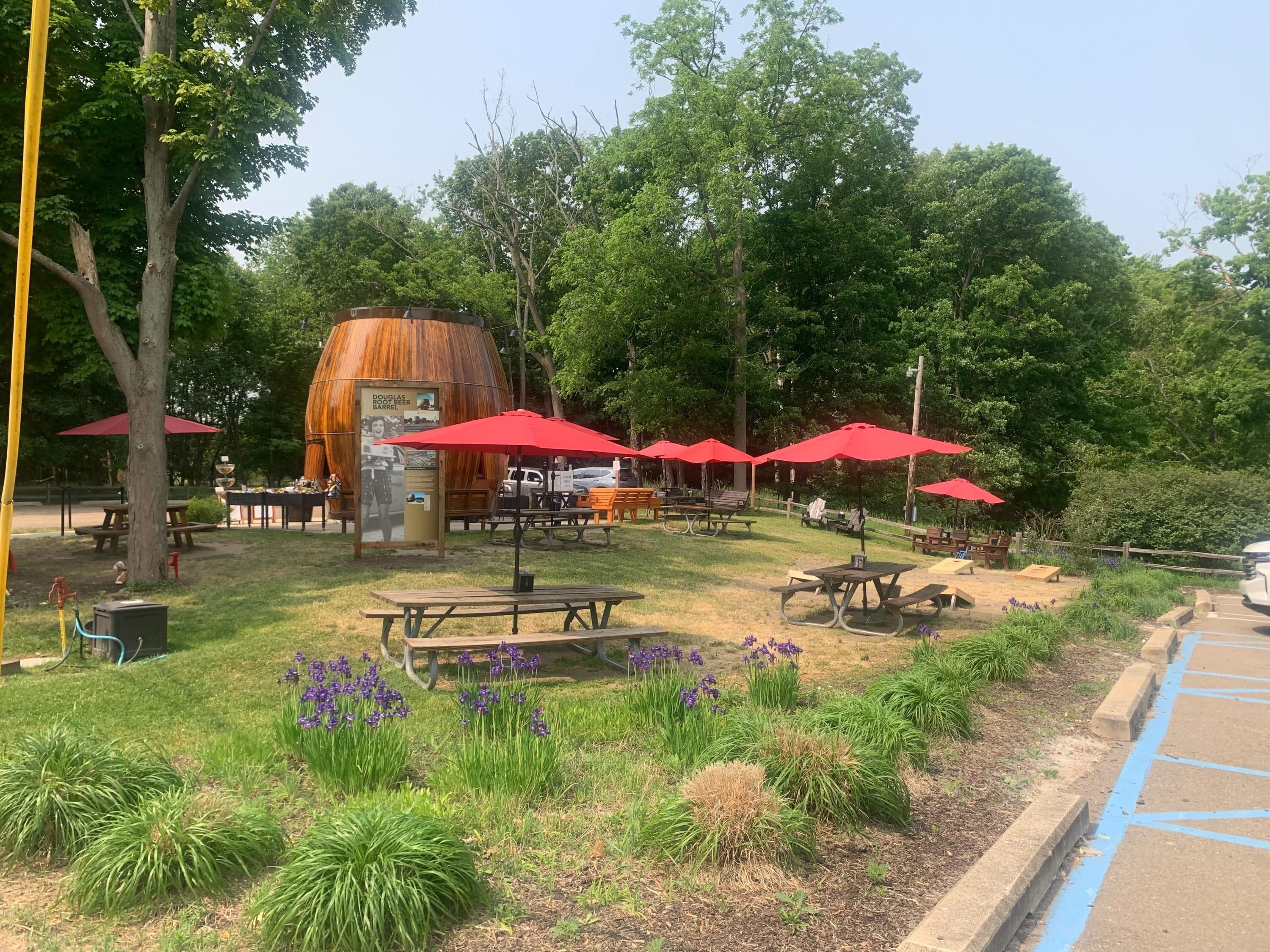 A park with picnic tables, red umbrellas, and a large barrel labeled 'Douglas Root Beer Barrel'.