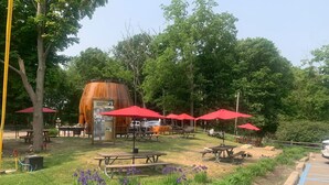 Un parque con mesas de picnic, sombrillas rojas y un barril grande con la leyenda 'Douglas Root Beer Barrel'.