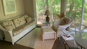 Una sala de estar acogedora con un sofá, una mesita con una lámpara y un sillón. Se ve una ventana con vistas a árboles.