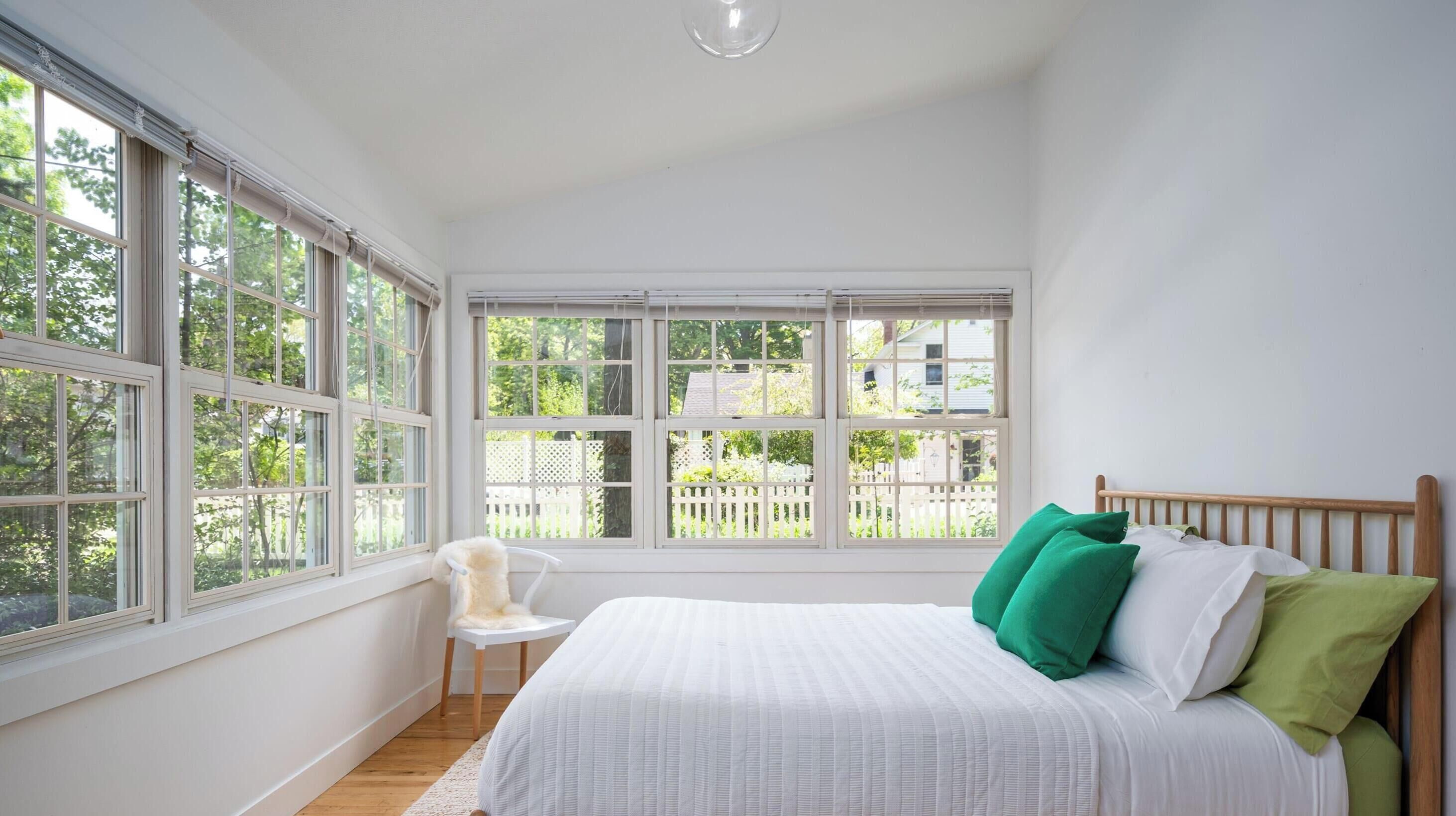 A bedroom with a wooden bed, white bedding, and green pillows. There is a chair and a large window with a view of greenery and a white fence.