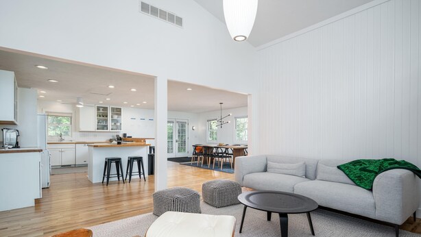 Un espace de vie moderne à aire ouverte, avec un canapé gris, des poufs et une table basse. On aperçoit une cuisine et une salle à manger à l’arrière-plan.