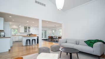 Un espacio habitable moderno y abierto con un sofá gris, otomanas y una mesita de centro. Se ve una cocina y zona de comedor al fondo.