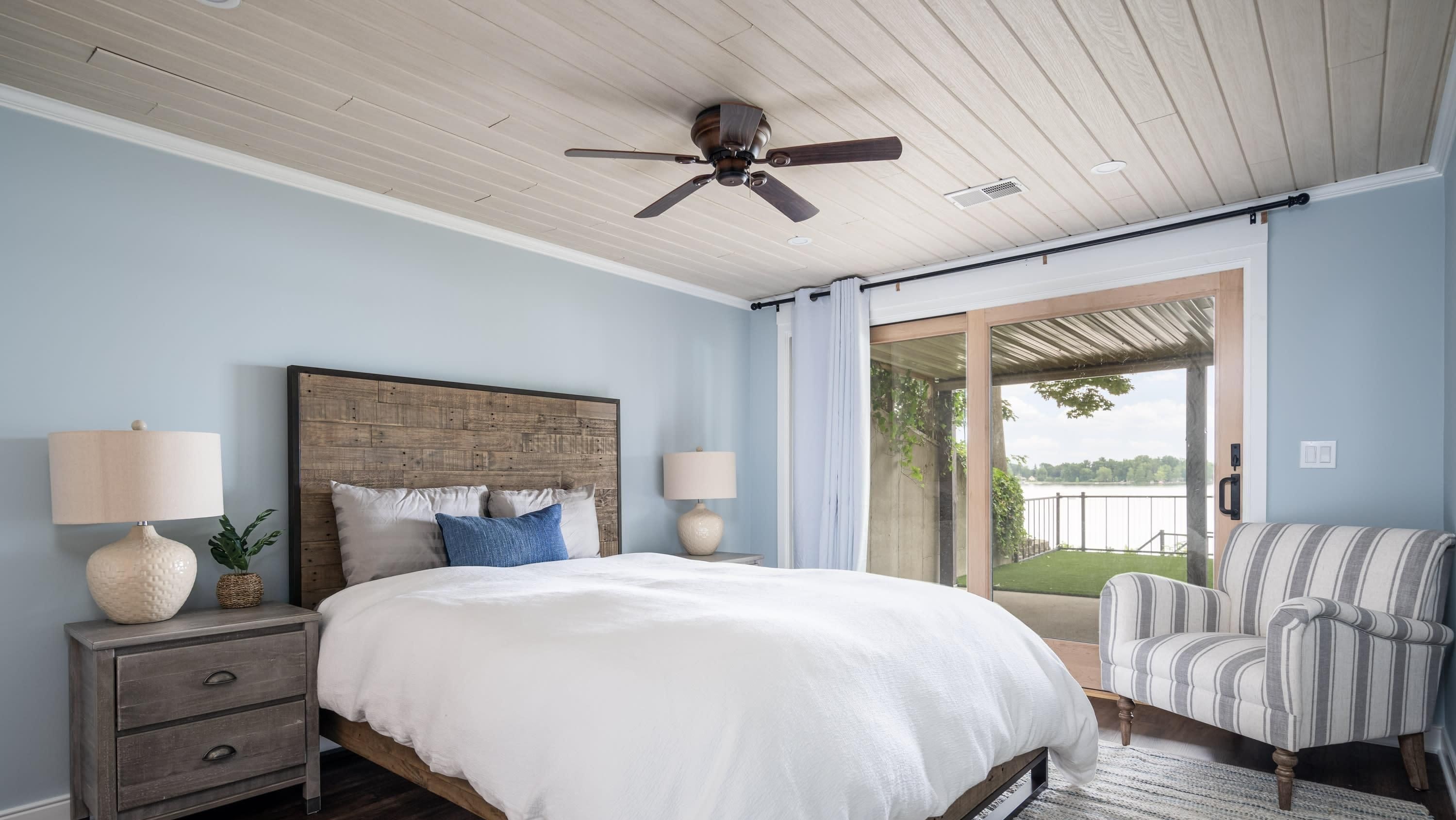 A bedroom with a wooden headboard, a large bed with white bedding, two bedside lamps, a striped armchair, and a view of a balcony with a railing.