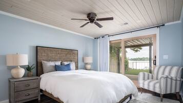 A bedroom with a wooden headboard, a large bed with white bedding, two bedside lamps, a striped armchair, and a view of a balcony with a railing.
