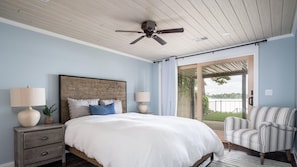 Un dormitorio con cabecera de madera, una cama grande con ropa de cama blanca, dos lámparas de noche, un sillón a rayas y vistas a un balcón con barandilla.