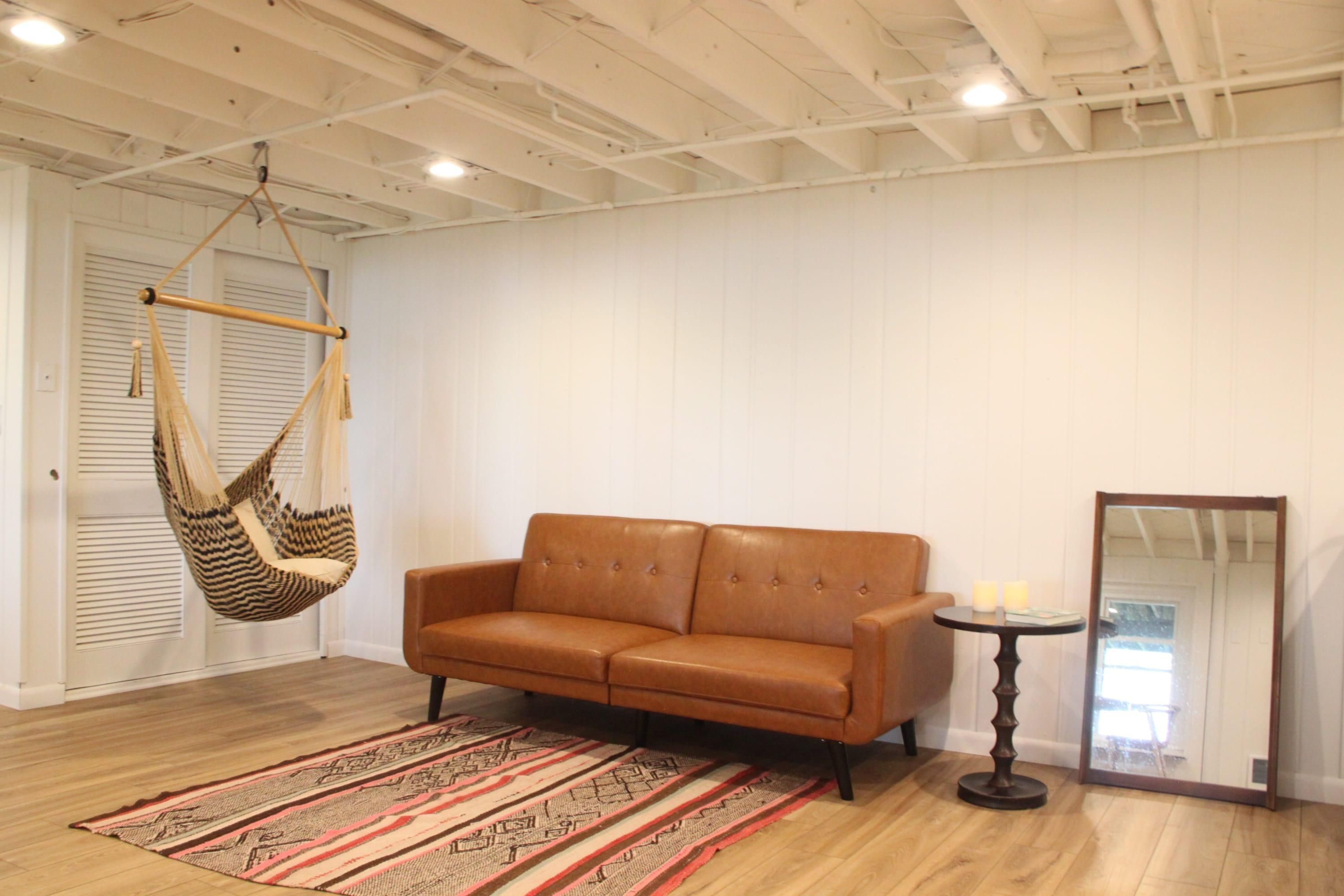 A room with a brown leather sofa, a hanging chair, a patterned rug, a round table with candles, and a large mirror.