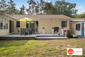 A covered patio area with outdoor furniture and a grill, surrounded by a grassy yard and trees.